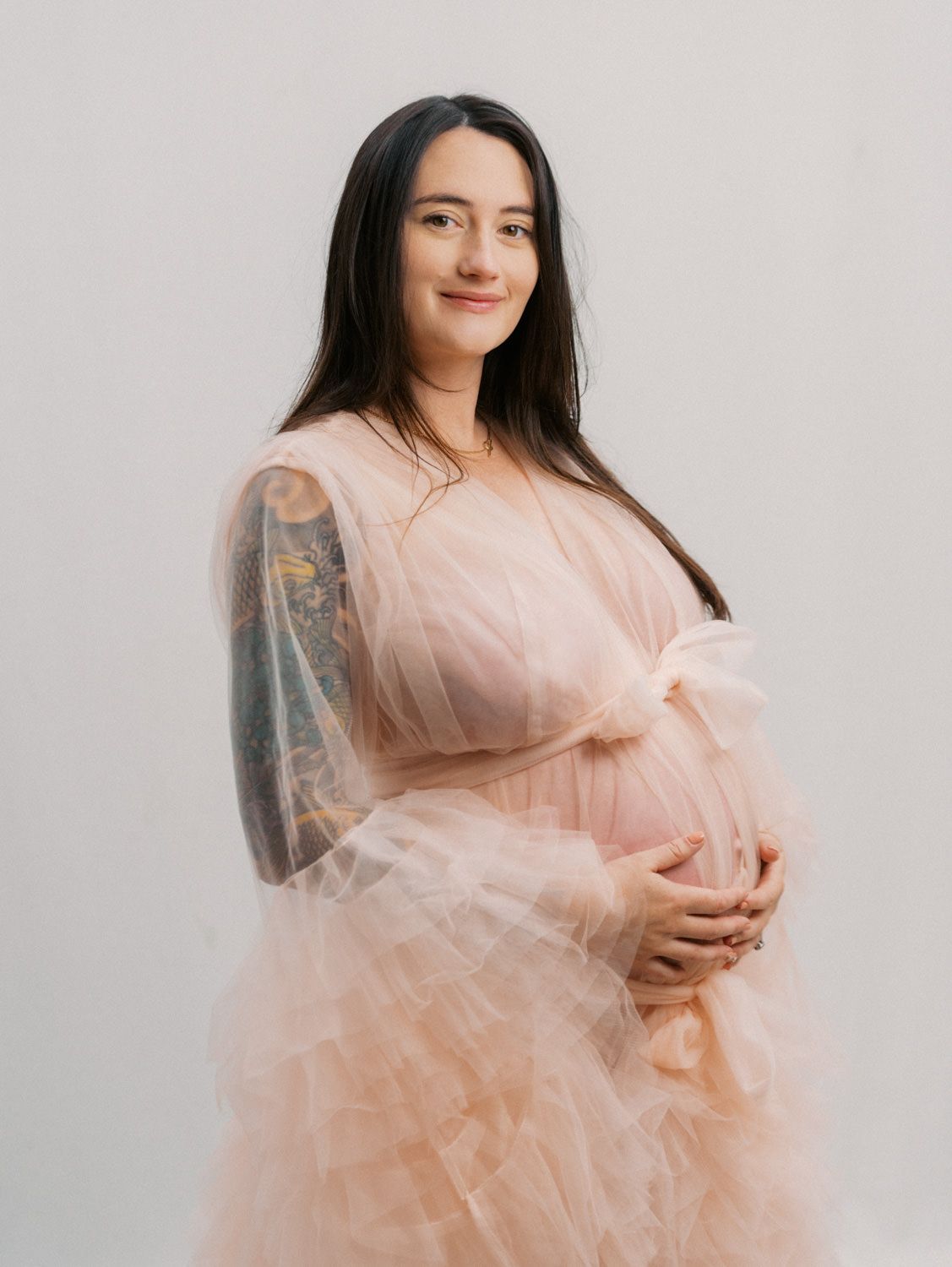 Pregnant woman in a blush pink tulle gown with sleeve tattoos cradling her baby bump in a studio maternity portrait.