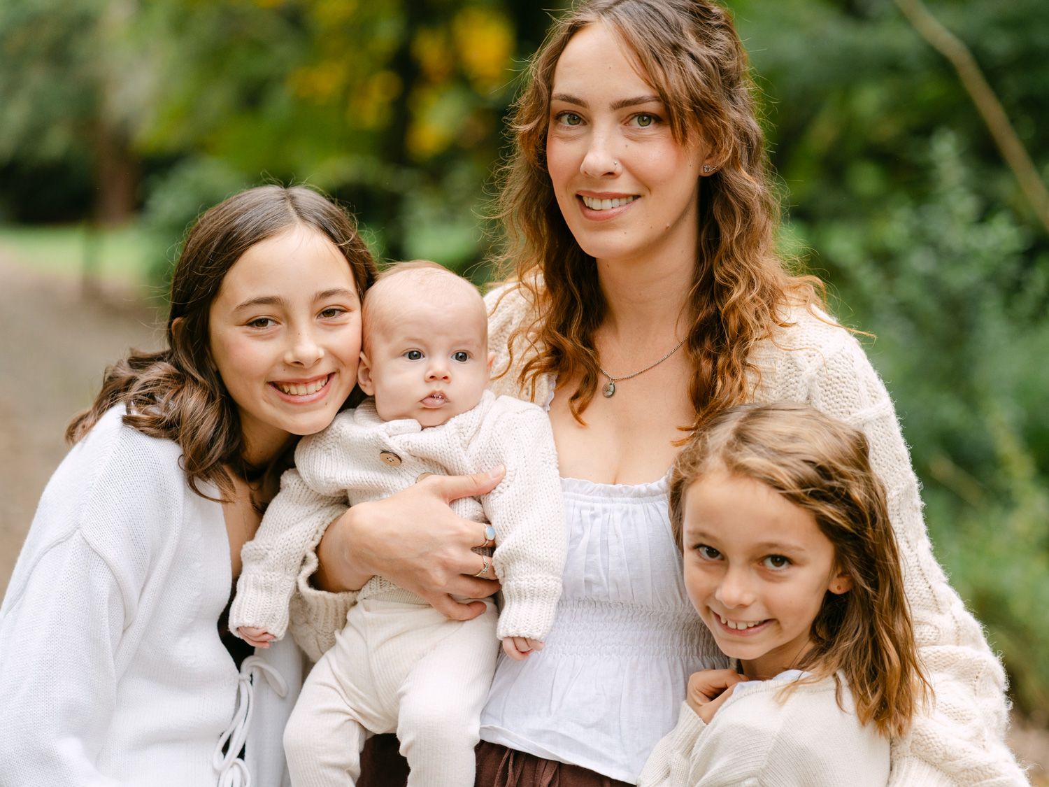 A young mother holds her baby surrounded by two daughters, all wearing white, smiling outdoors in a lush green setting.