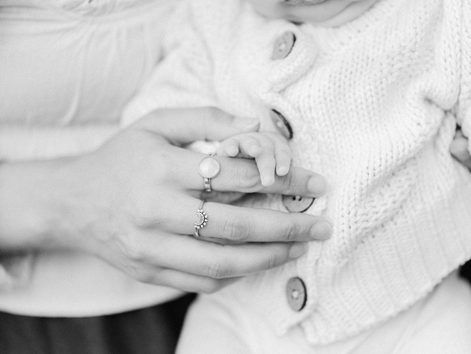Close-up black and white photo of adult hands cradling a newborn baby's tiny hand, with a delicate ring visible.