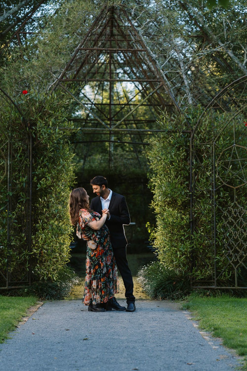 Couple embracing on a garden path beneath a rustic metal pyramid arch surrounded by lush green hedges.