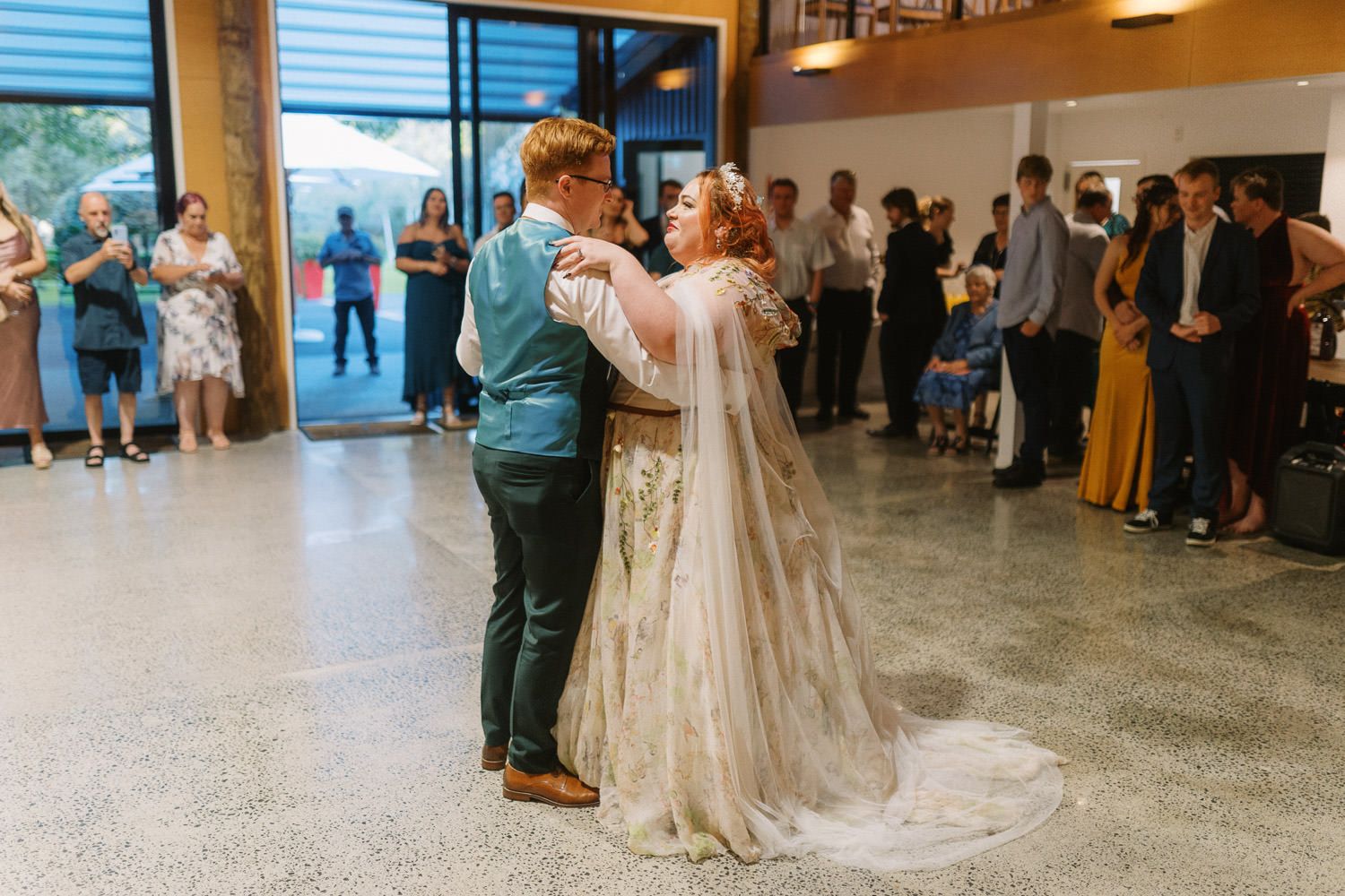 Bride and groom share their first dance at wedding reception with guests watching in elegant venue.