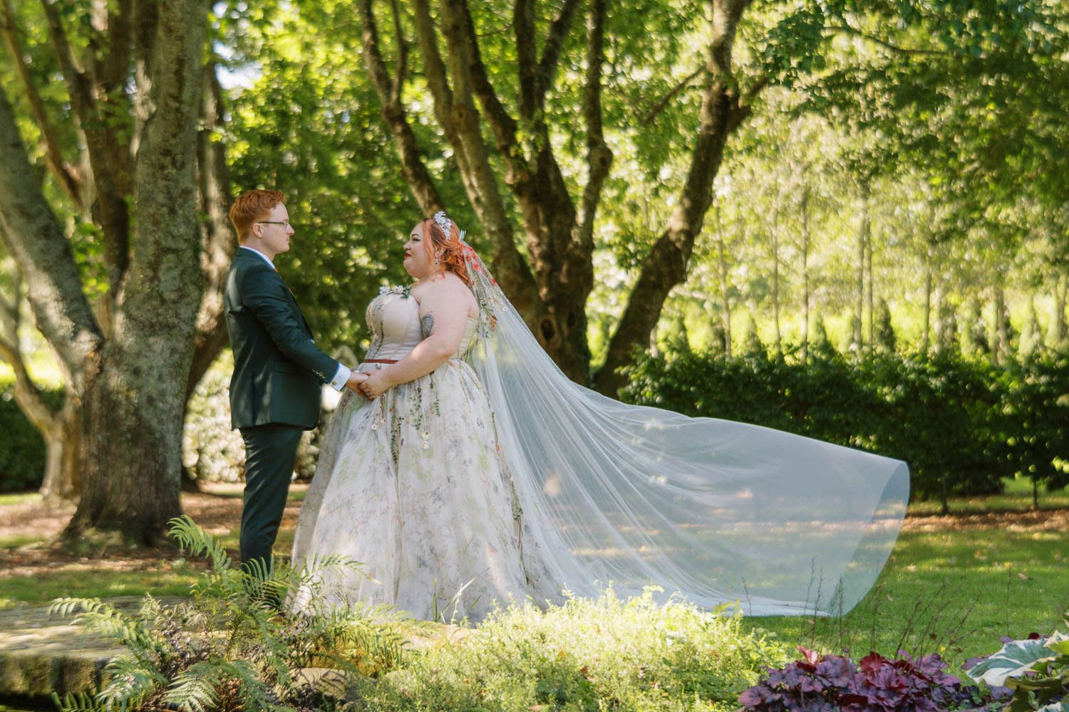 Bride and groom share a romantic moment outdoors, her floral gown and flowing veil catching the breeze beneath lush green trees.