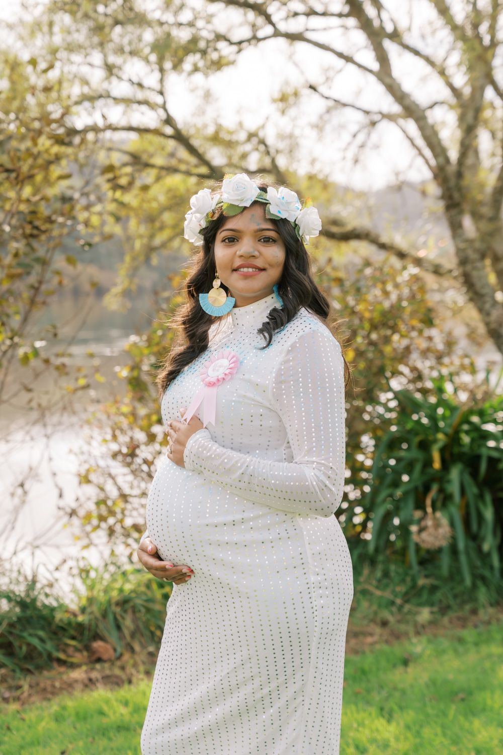 Pregnant woman in white crochet dress and floral crown holds baby items outdoors near a serene lake.