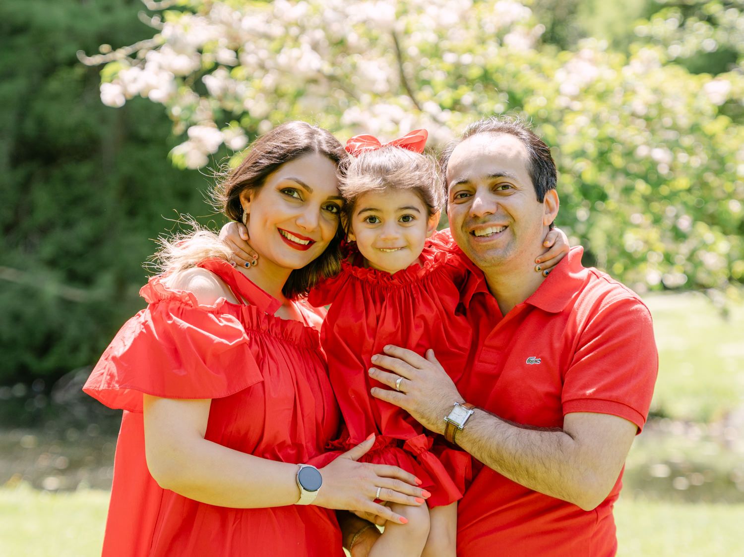 Happy family of three wearing matching red outfits posing outdoors with blooming trees in background.