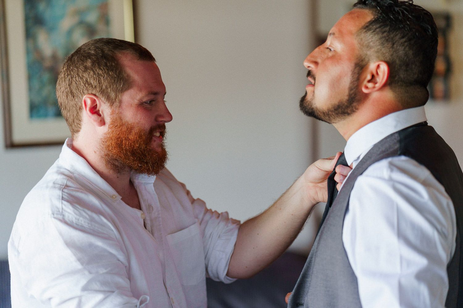 A bearded groomsman adjusts the bow tie of a groom in a gray vest before a wedding ceremony.
