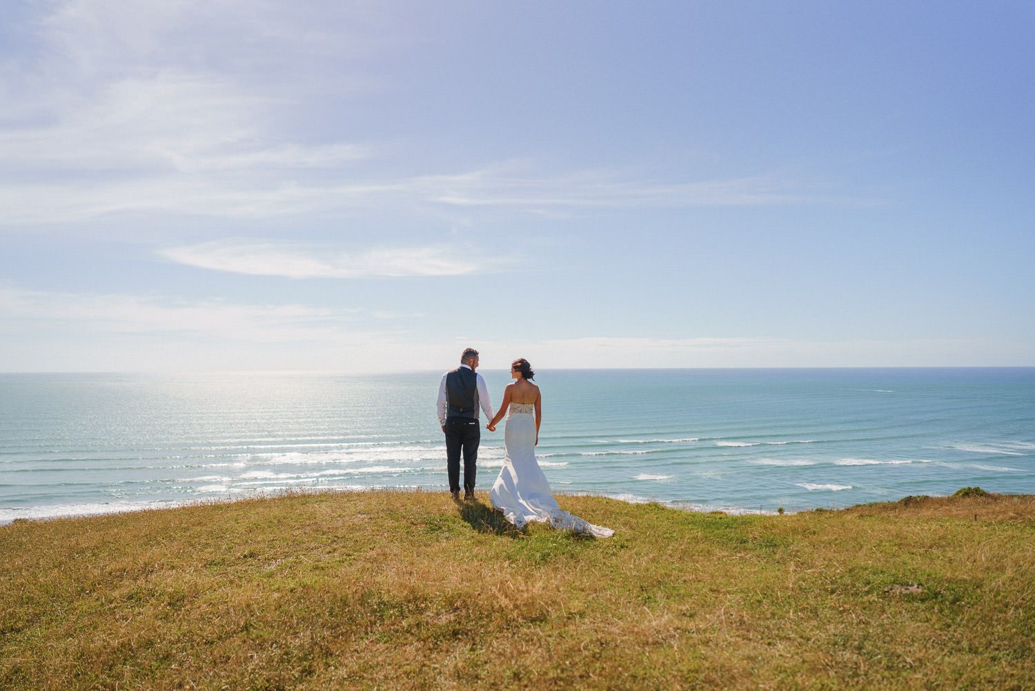 Bride and groom holding hands on a coastal cliff overlooking a turquoise ocean under a bright blue sky.