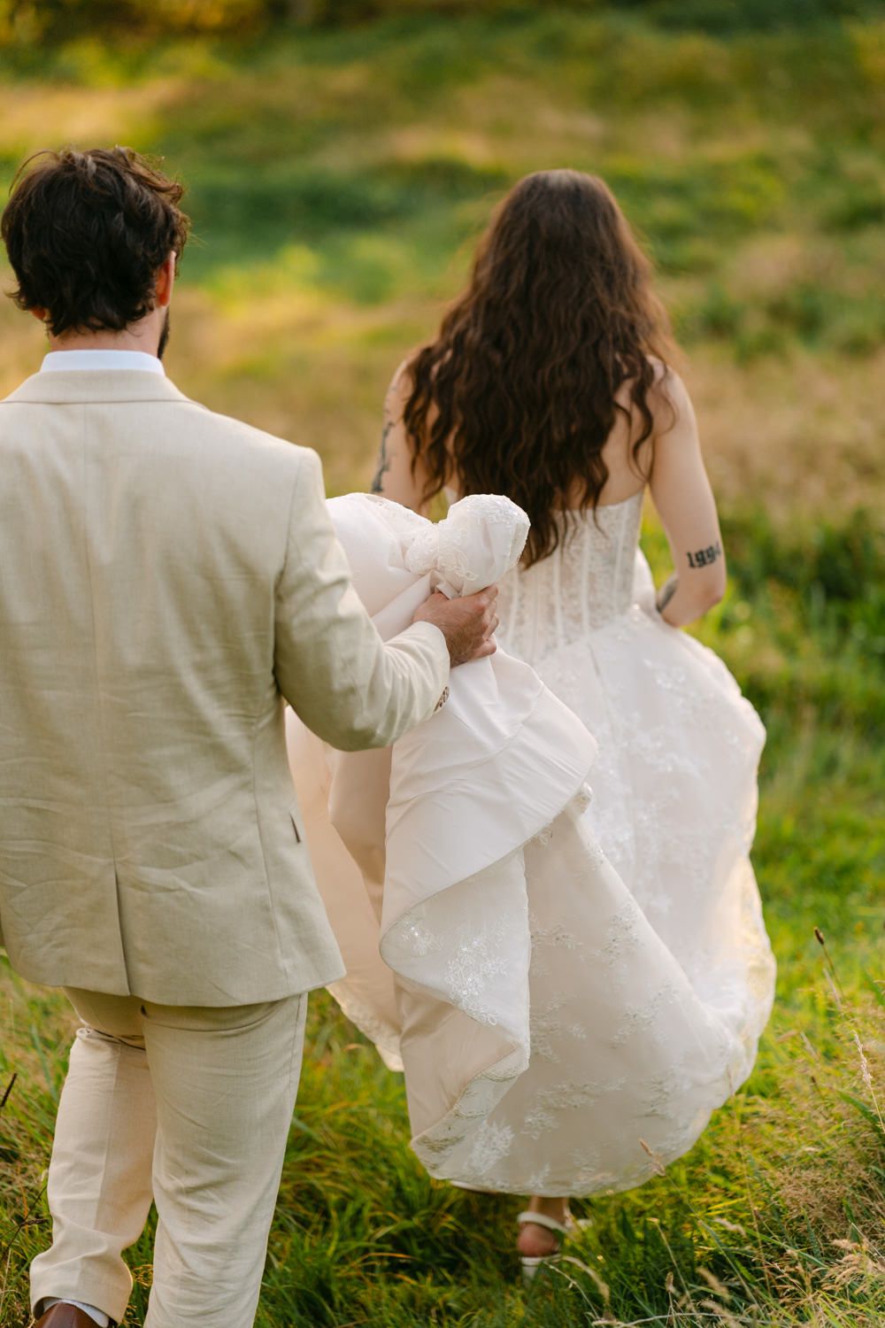 Bride and groom walking through green meadow, viewed from behind, bride in white lace gown, groom in cream suit.
