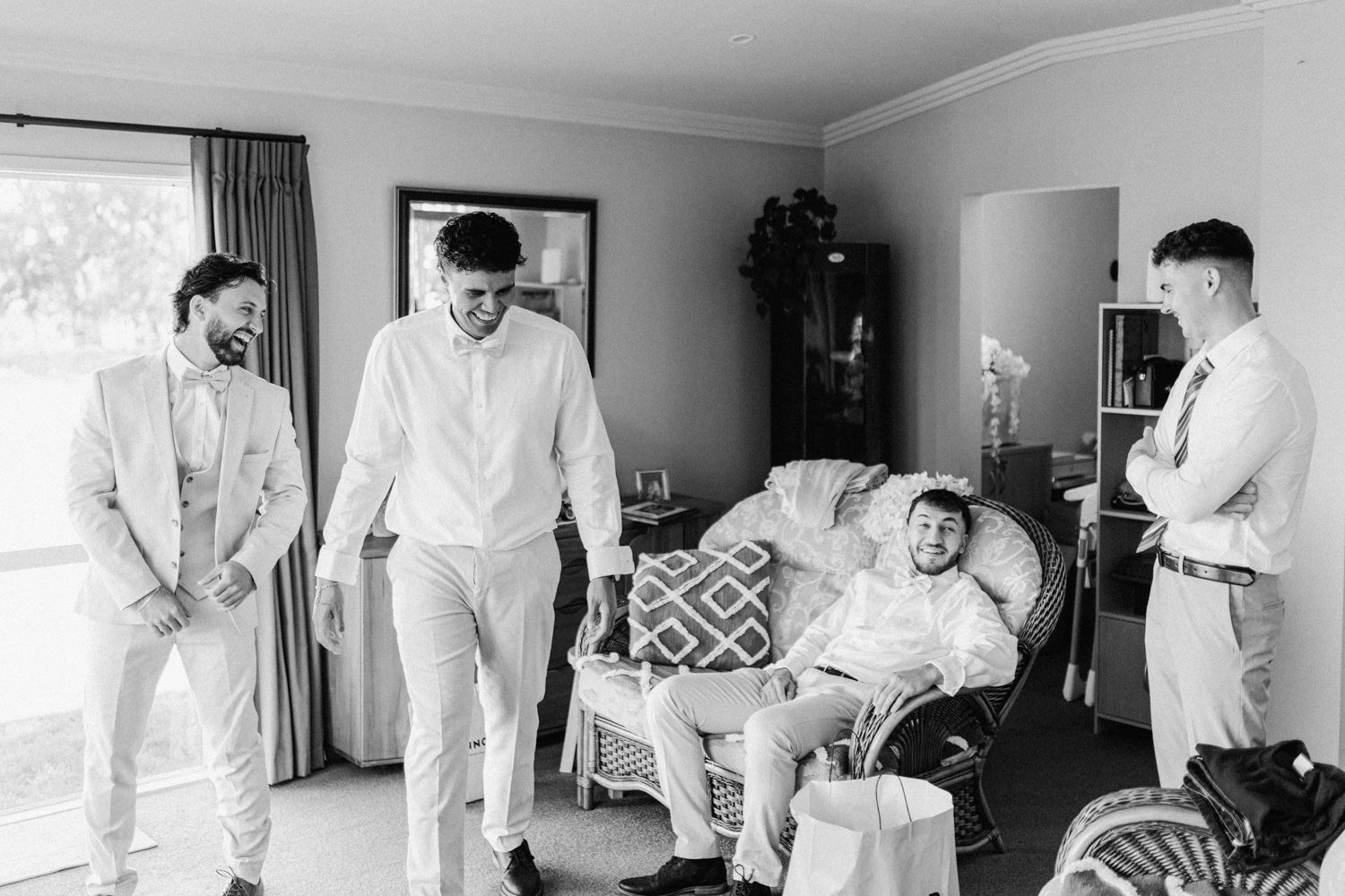 Groomsmen laughing and relaxing together in a room during wedding preparations, captured in black and white.