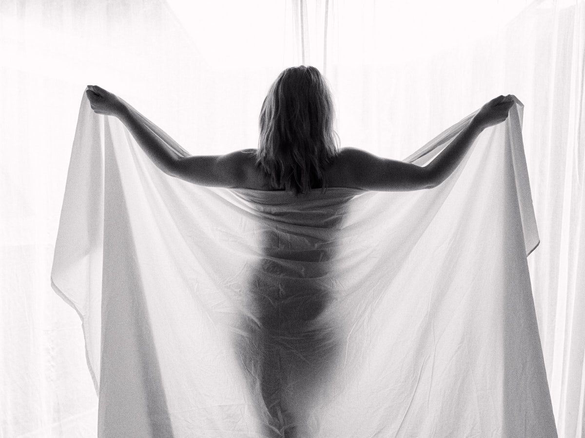 Black and white artistic photo of a woman holding a white sheer fabric sheet behind her back.