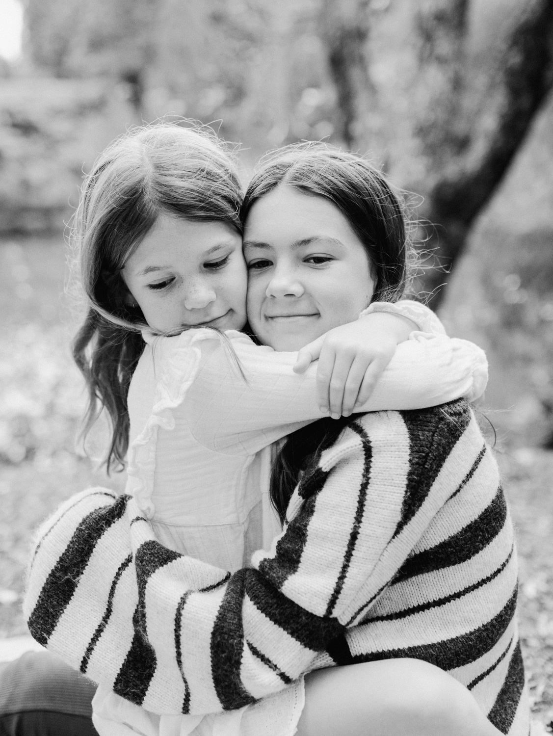 Two young girls share a sweet embrace outdoors in a black and white portrait photo.