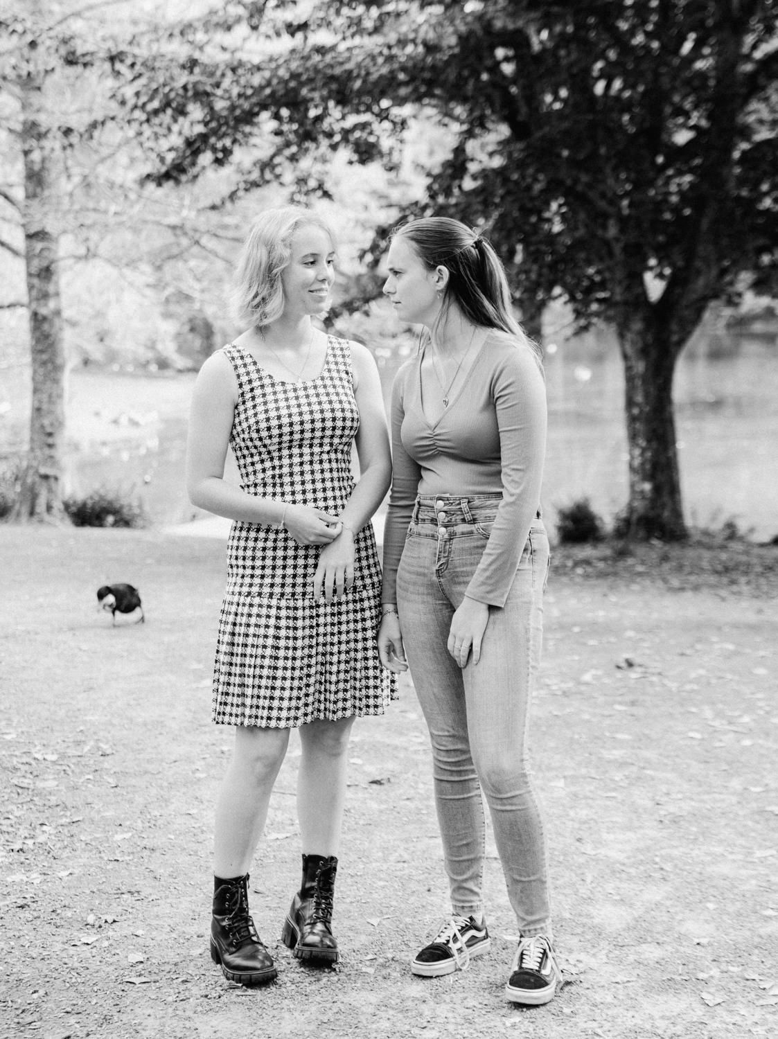 Two young women smiling at each other in black and white outdoor photo near trees.