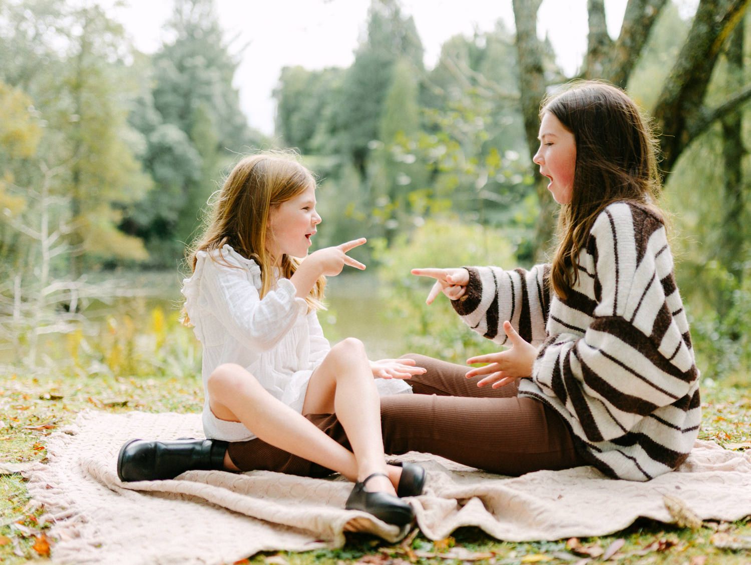 Two girls sitting on a blanket outdoors, laughing and pointing at each other in a park setting.