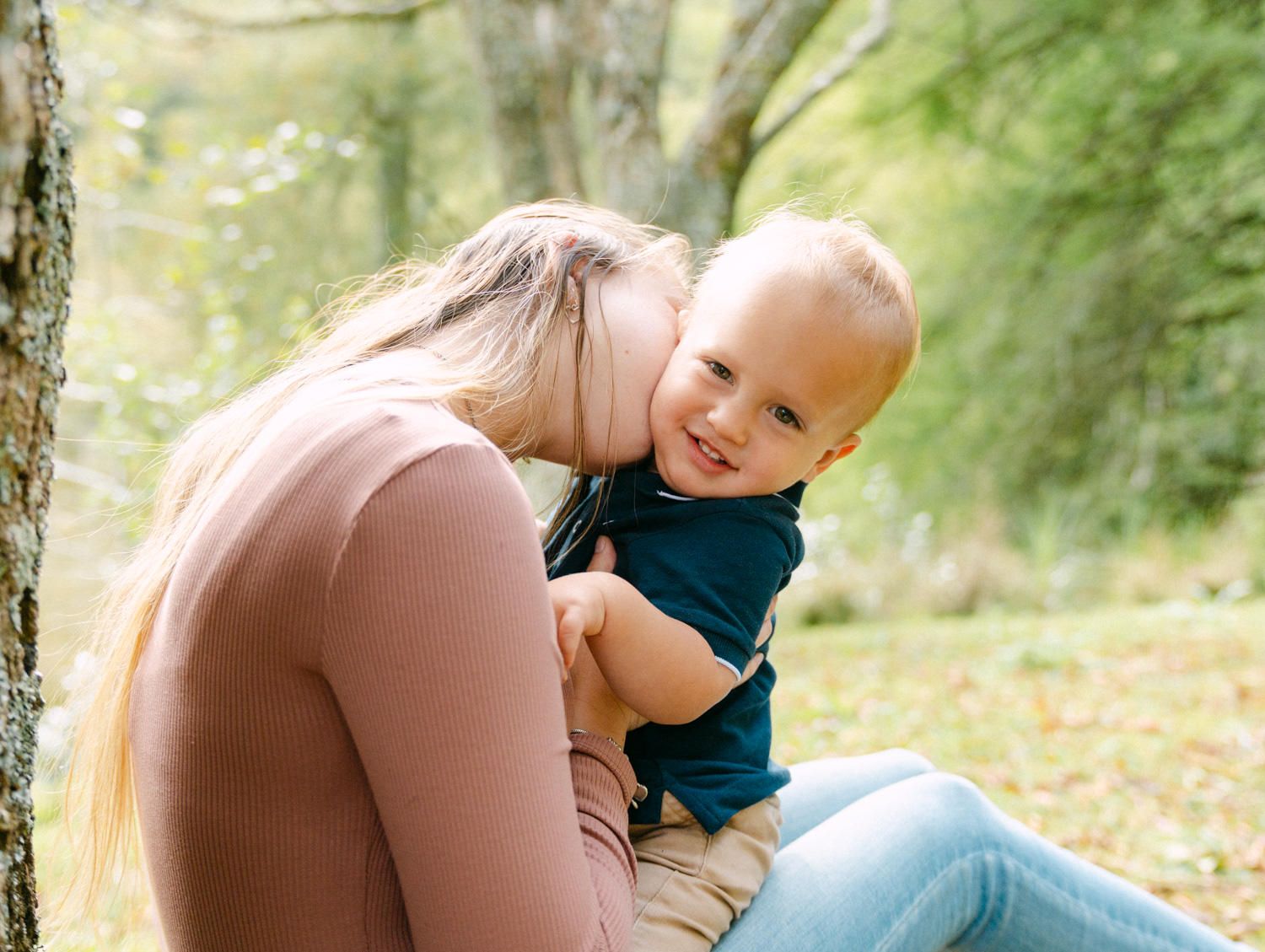 A mother tenderly kisses her smiling baby boy outdoors in a lush green park setting.