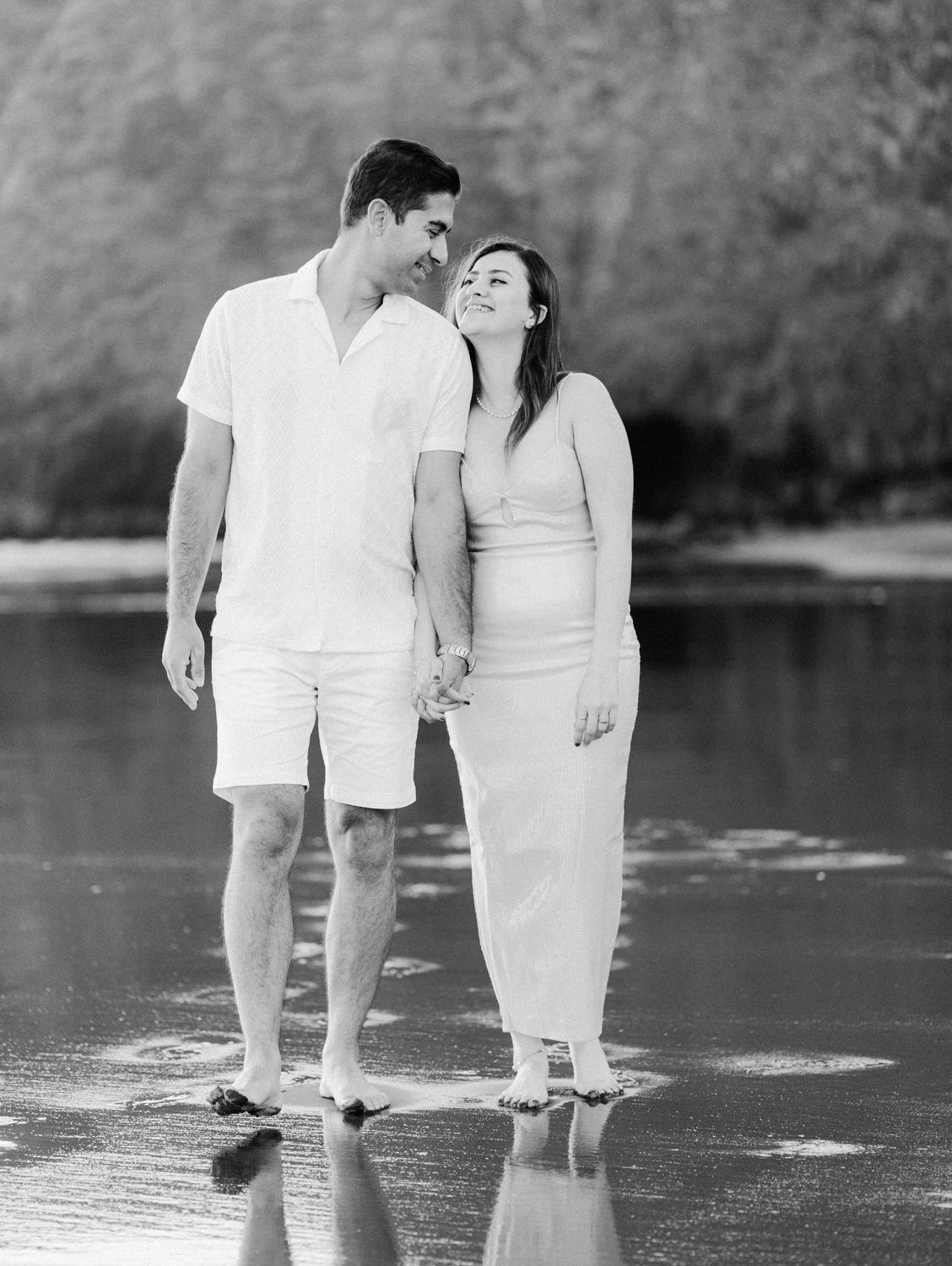 Black and white photo of a couple holding hands and walking barefoot on a reflective beach shore.