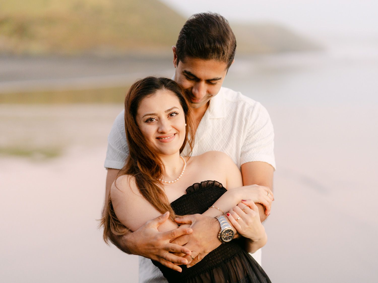 Couple embracing on a beach at sunset, man in white shirt hugging woman in black dress, romantic outdoor photo.