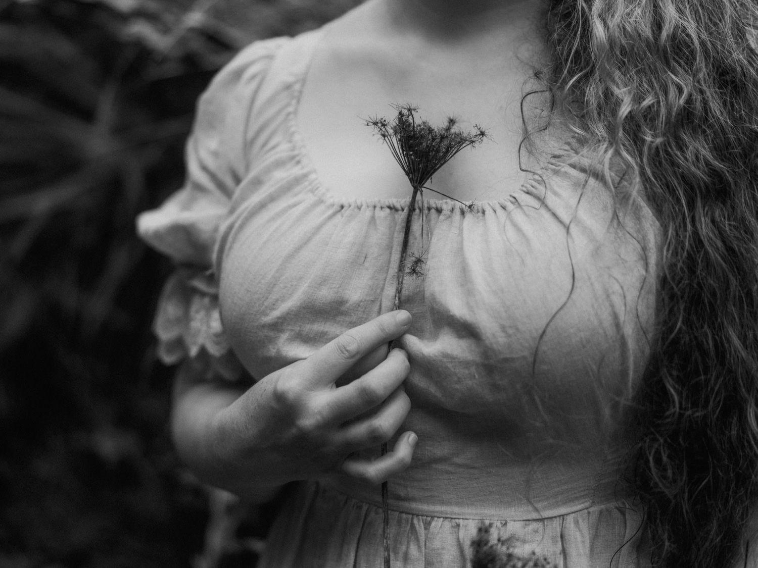 Close-up of woman in vintage white dress holding dried wildflower in black and white photo.