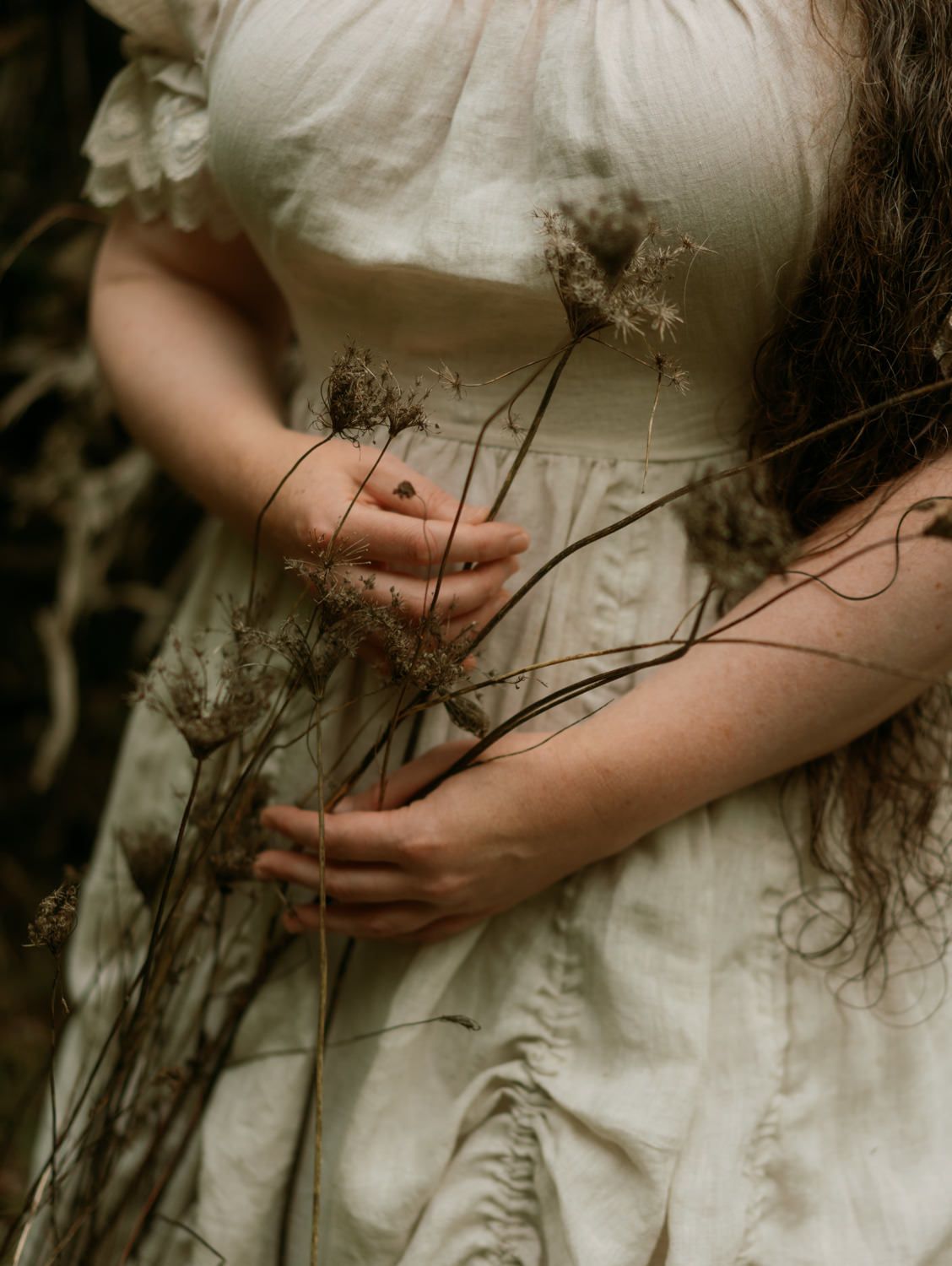 Woman in white linen dress holding dried wildflowers, moody nature photography with dark forest background.