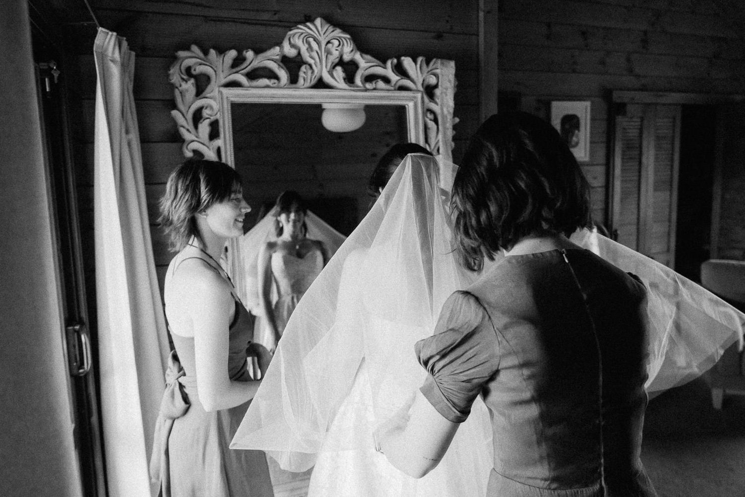 Black and white photo of a bride having her veil adjusted by a bridesmaid in front of an ornate mirror.