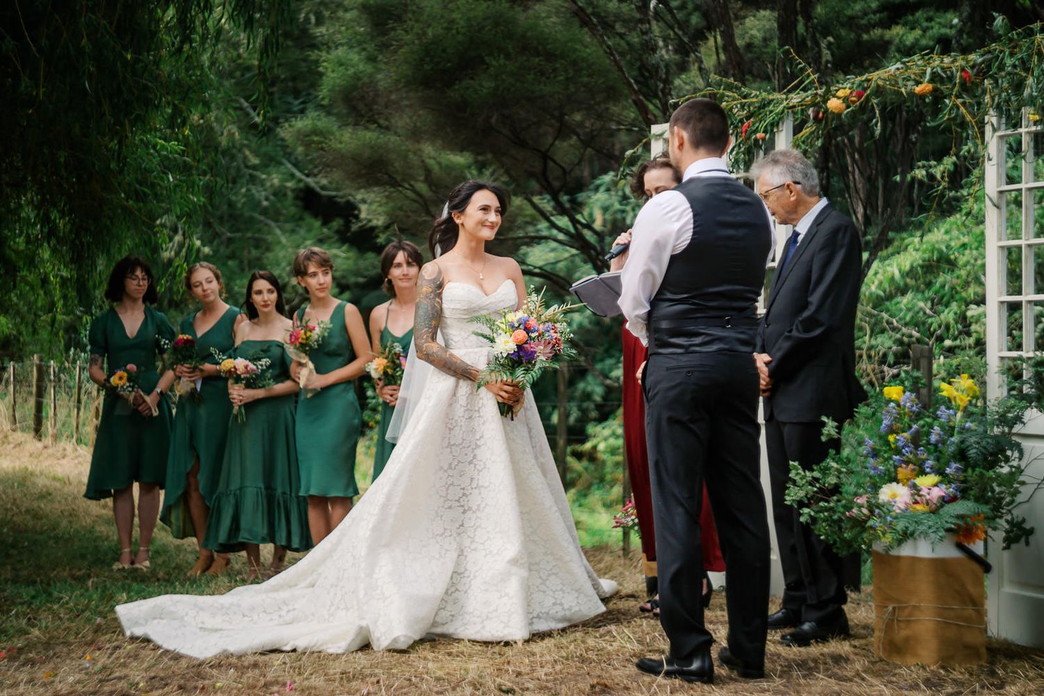 Bride in white lace gown smiles at groom during outdoor wedding ceremony with bridesmaids in green dresses.