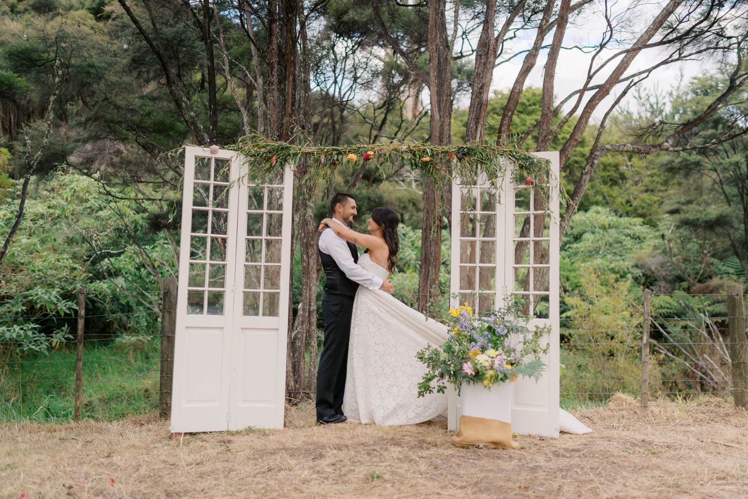 Couple sharing romantic kiss at outdoor wedding ceremony with white vintage doors and lush greenery backdrop.