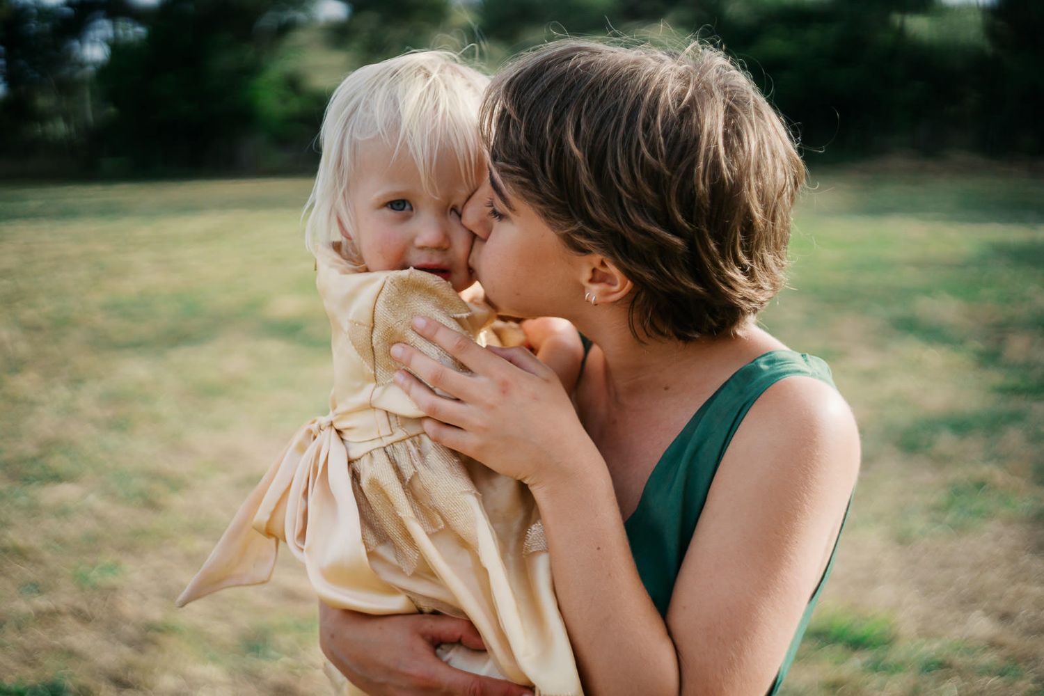 A mother tenderly kisses her smiling toddler daughter outdoors in a grassy field.