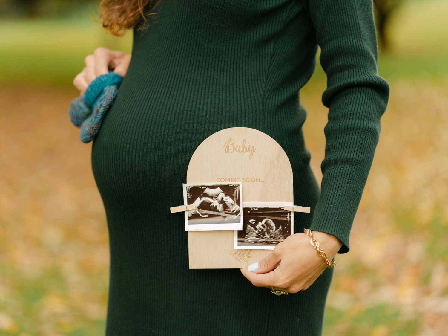 Pregnant woman in green dress holding ultrasound photos in wooden baby frame outdoors in autumn.