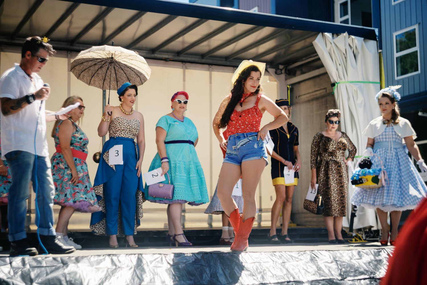 Women in vintage-style outfits pose on an outdoor stage at a retro fashion show on a sunny day.