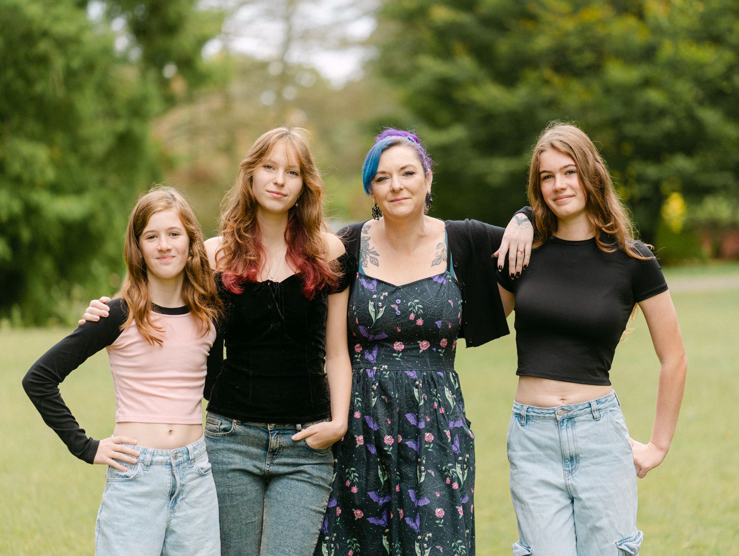 Four young women posing together outdoors on green grass, smiling in casual summer outfits.