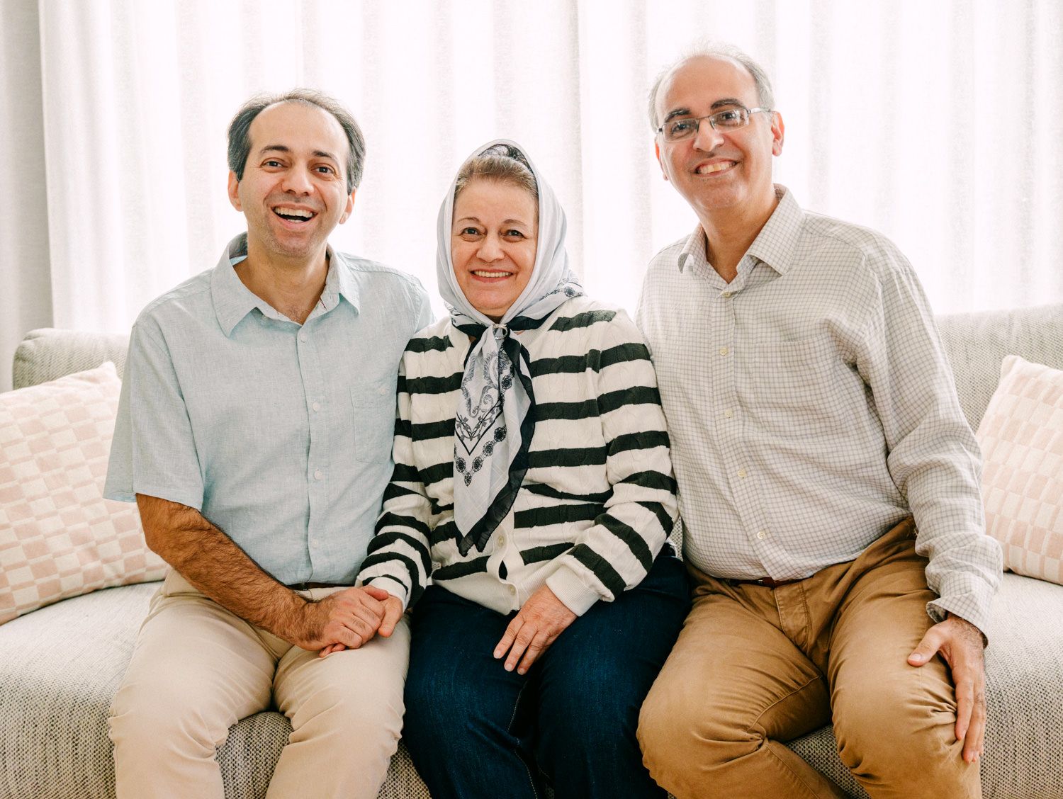 Three smiling adults sitting together on a white sofa in a bright living room, posing for a family portrait.