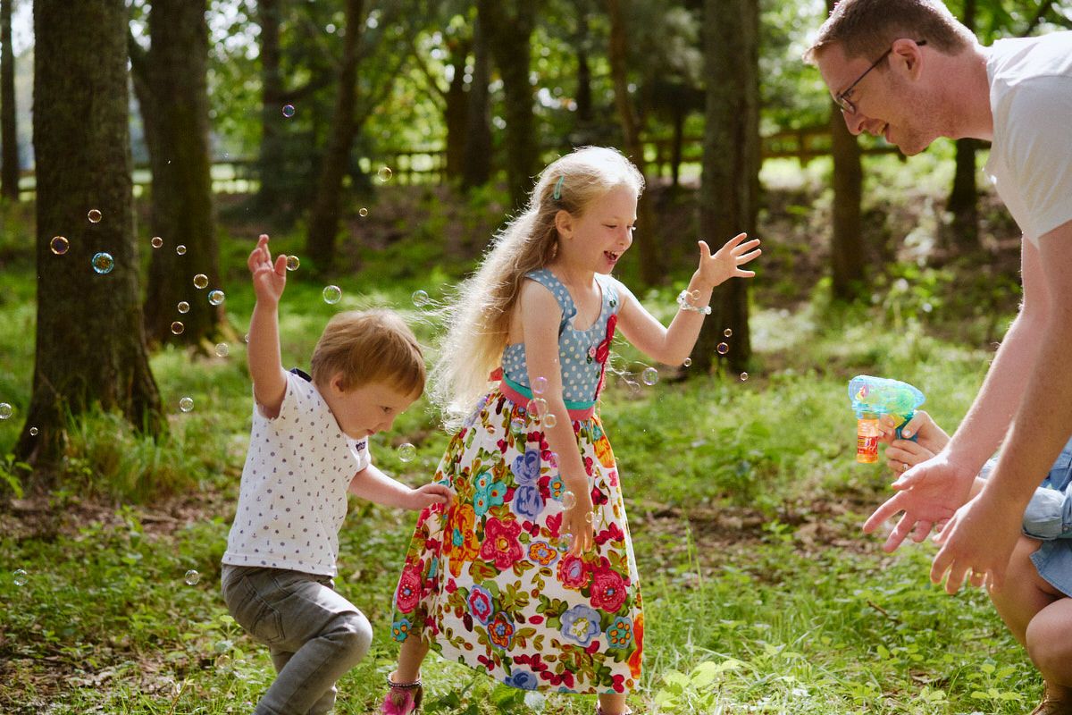 Two children and a man joyfully chasing bubbles in a sunny forest clearing, girl in floral dress, boy in white shirt.