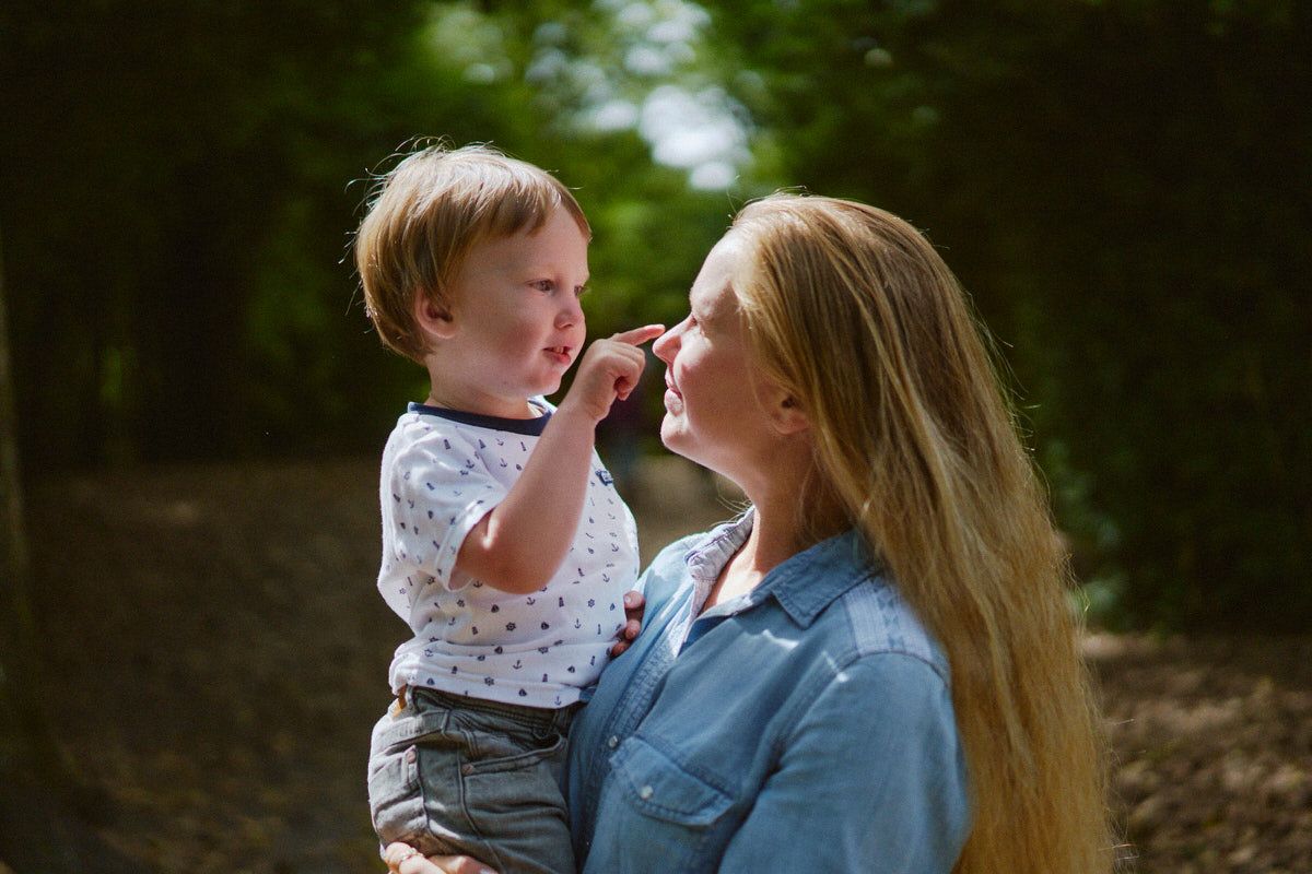 A mother holds her toddler son outdoors in a lush green forest, as the child gently touches her face.