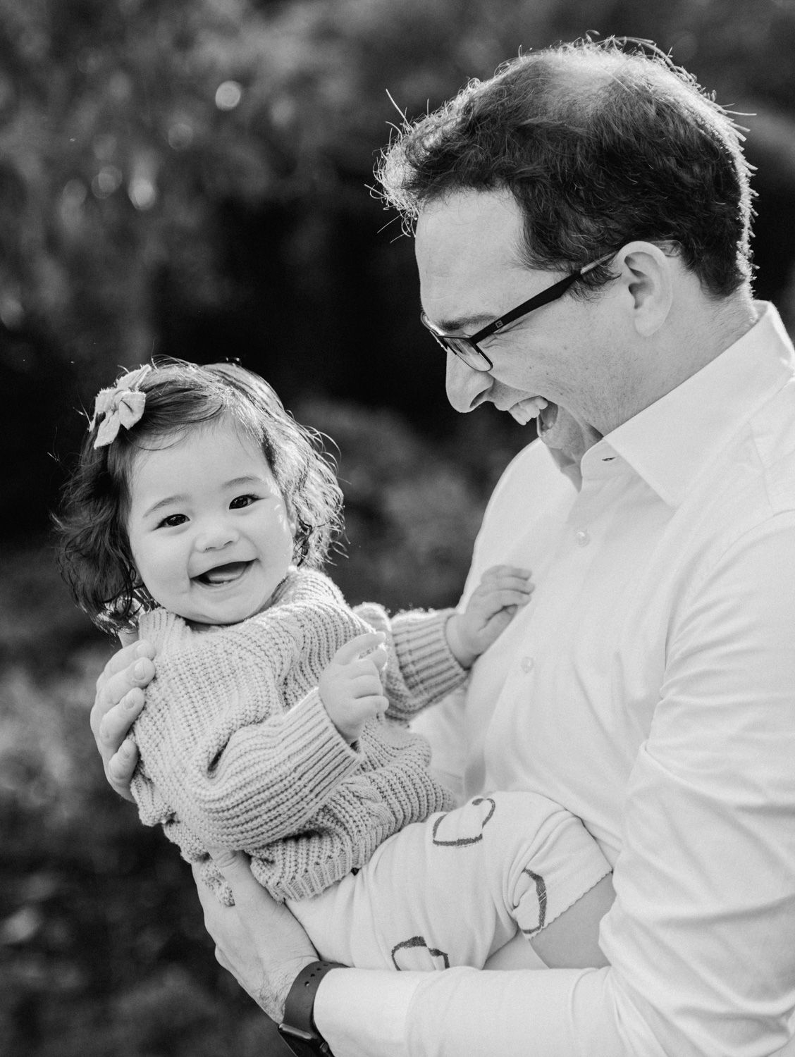 Black and white photo of a smiling man in glasses holding a happy baby girl in a knit sweater outdoors.