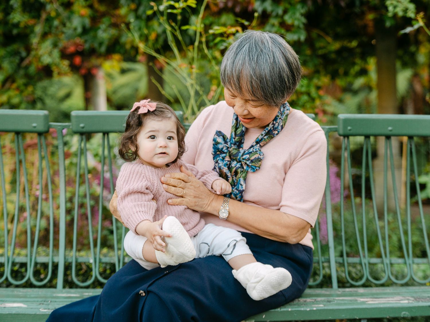 Grandmother sitting on a green park bench lovingly holding a baby girl in a pink sweater outdoors.