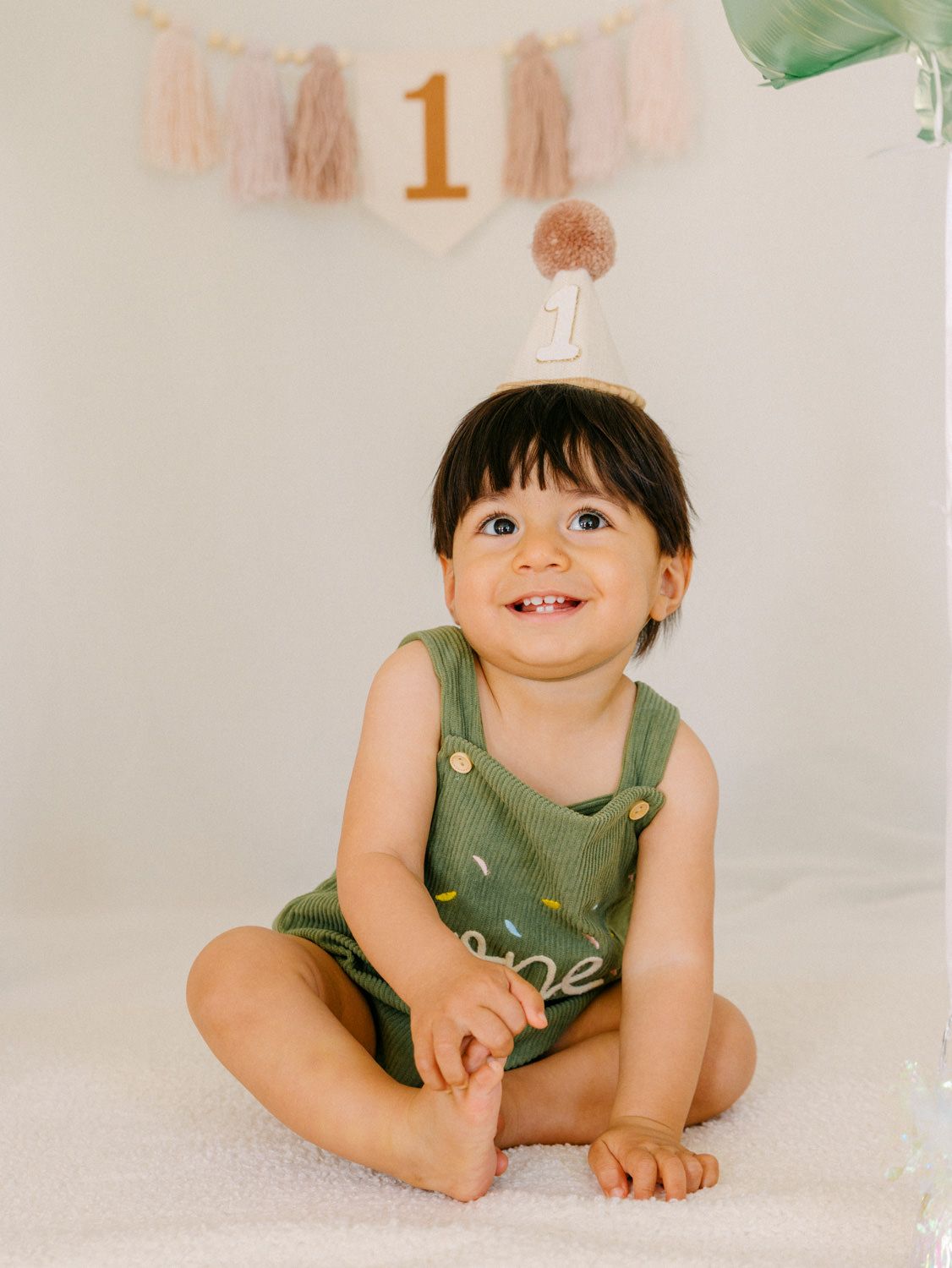 Smiling baby in green romper and party hat sits on white blanket at first birthday celebration.