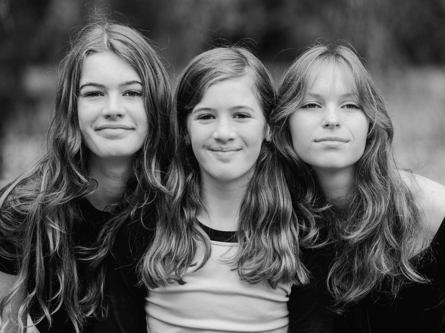 Black and white portrait of three smiling teenage girls with long hair posing closely together outdoors.