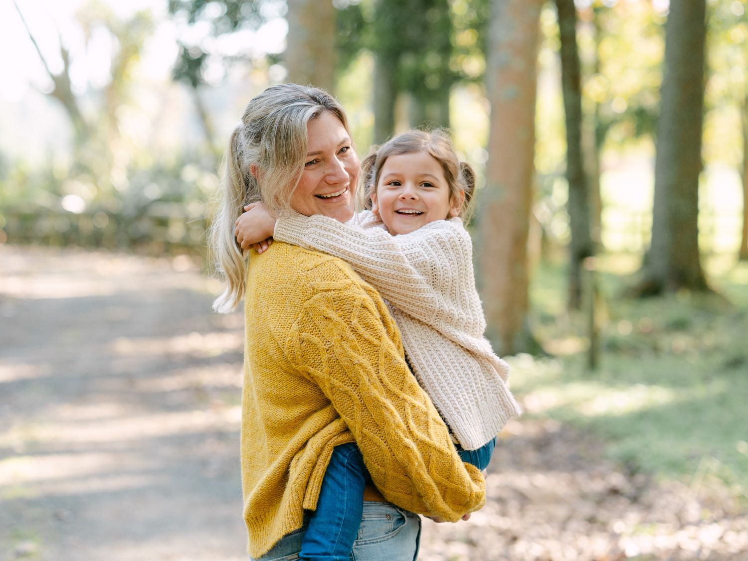 A smiling mother in a yellow sweater holds her young daughter in a beige knit sweater in a sunny forest path.