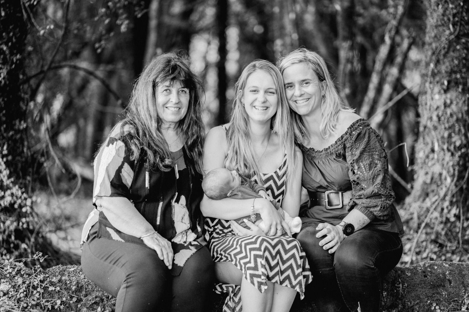 Three smiling women pose together outdoors in a forest setting in a black and white portrait photo.