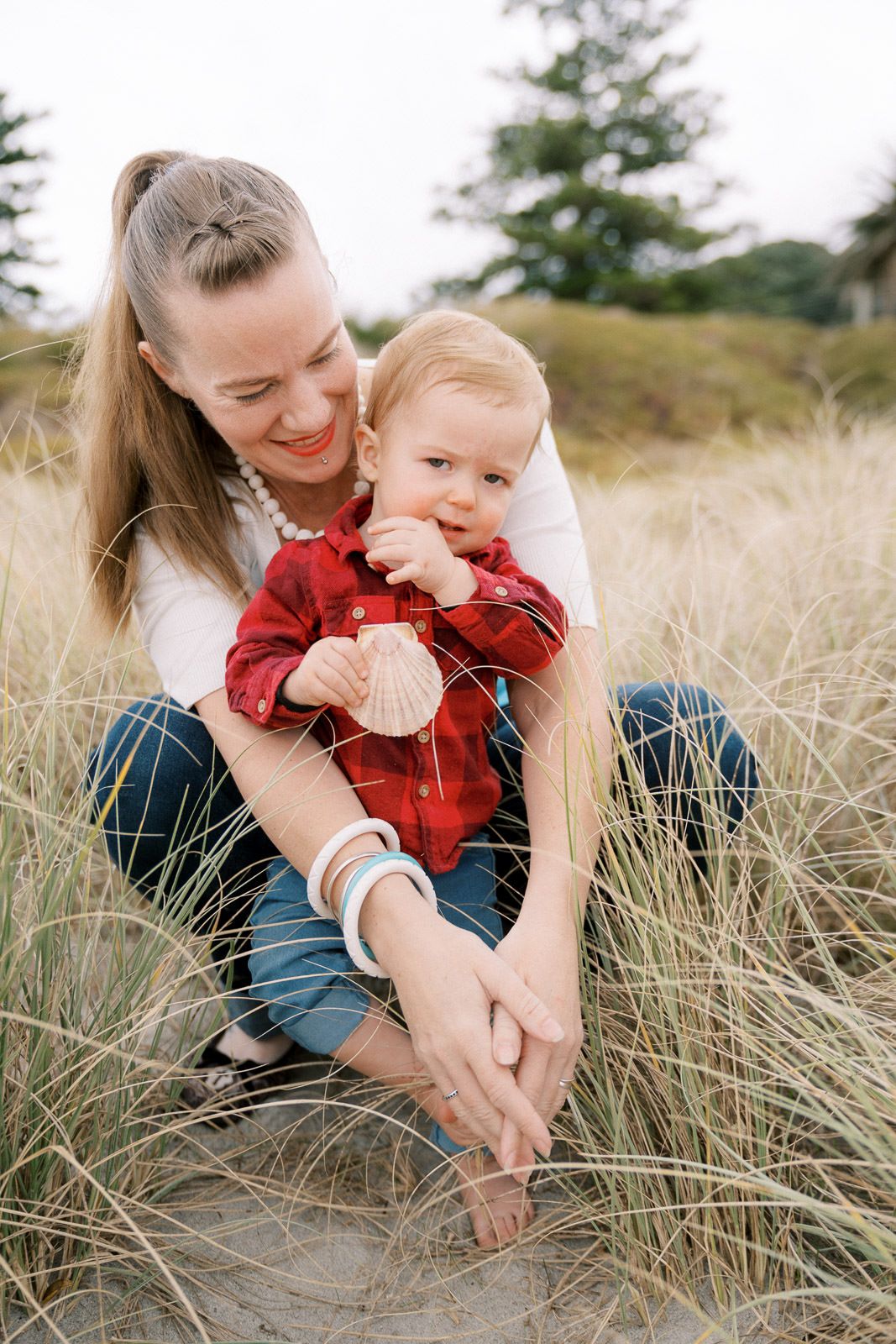 Smiling mother holding toddler in red plaid shirt exploring seashell in coastal beach grass.