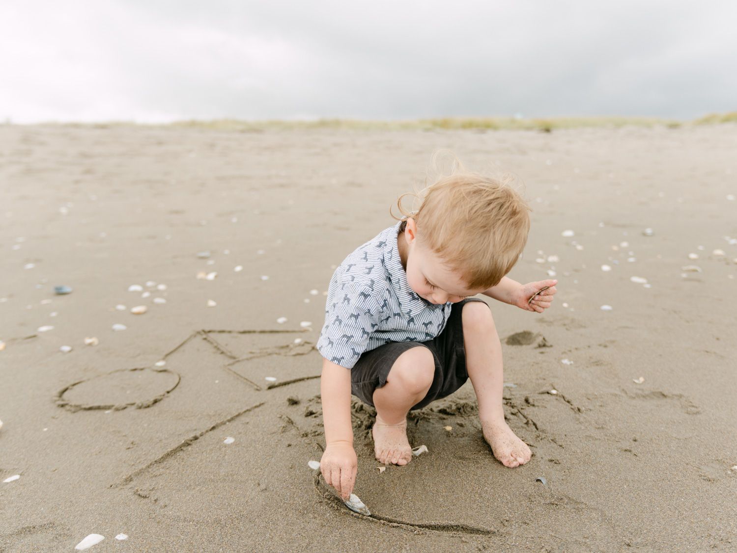 Toddler drawing in wet sand on a cloudy beach day, wearing a patterned shirt and dark shorts.