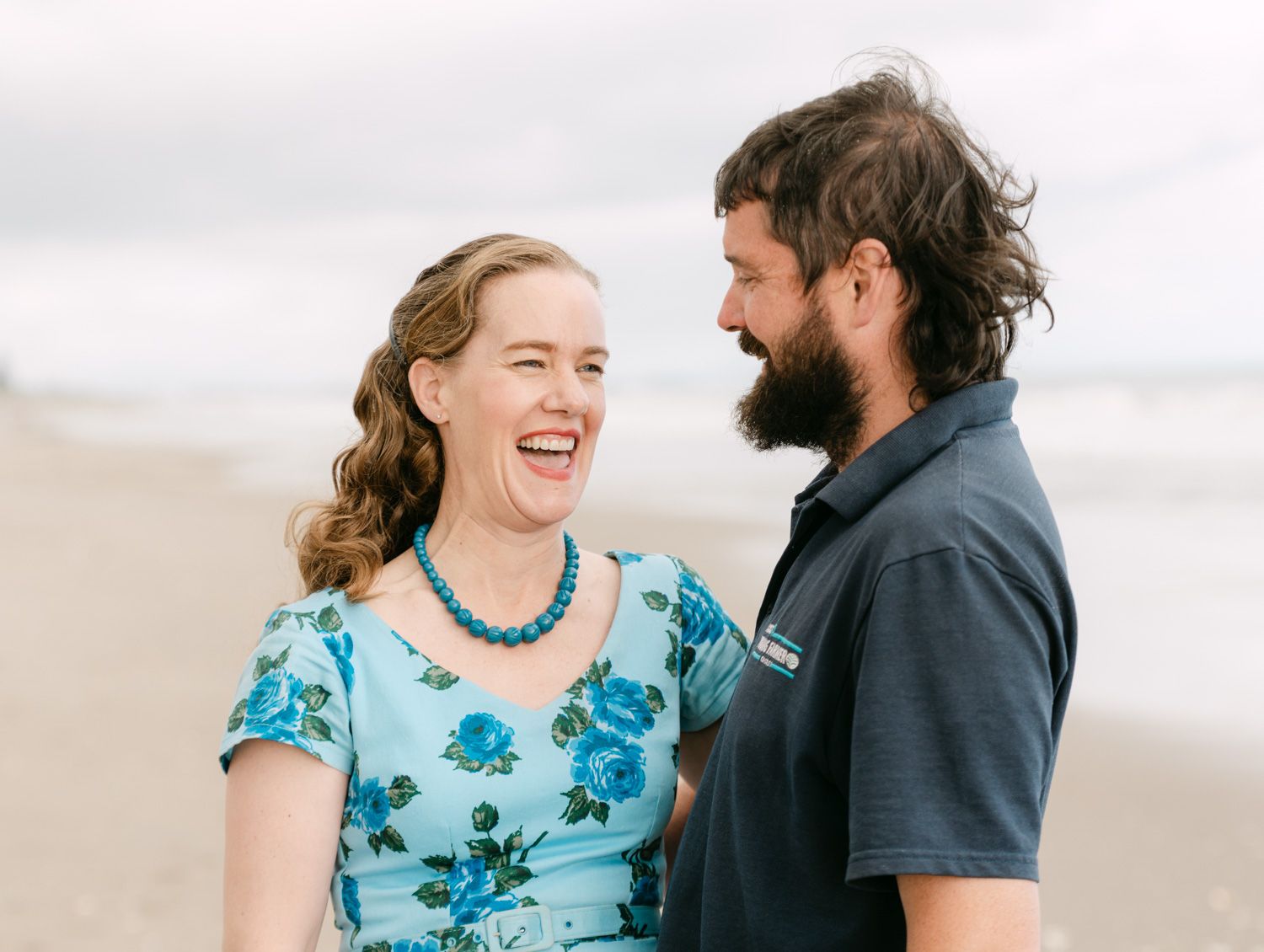 Couple laughing together on a beach, woman in blue floral dress with turquoise necklace, man with beard in gray polo.