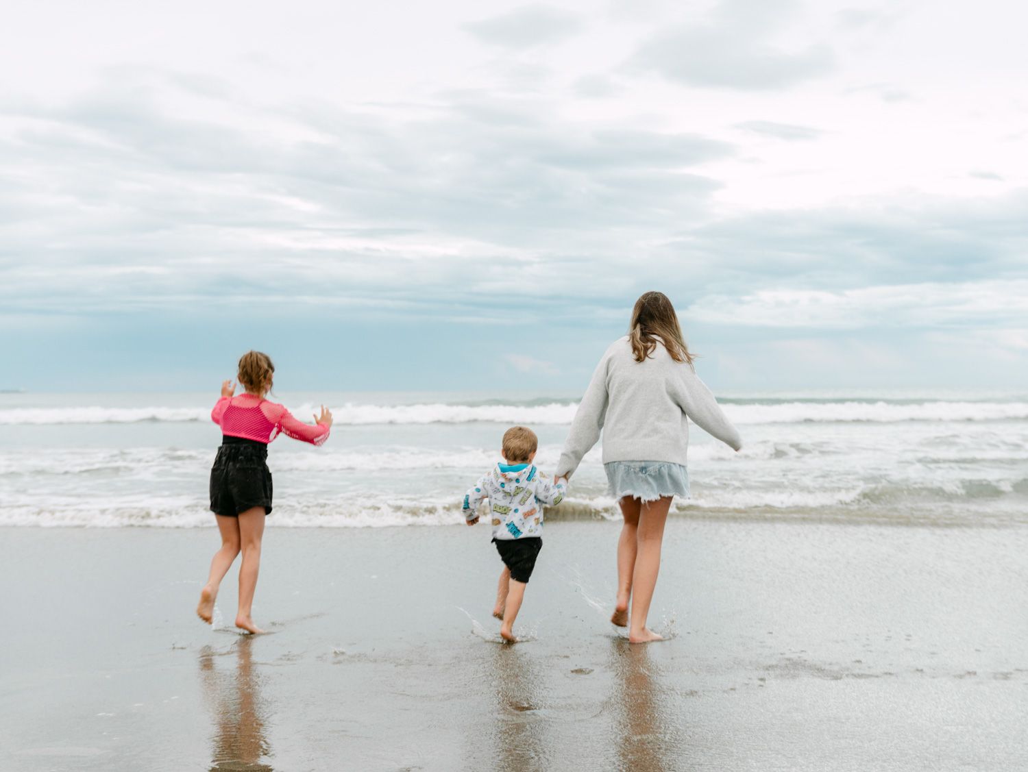 Three children walking hand-in-hand toward ocean waves on a cloudy beach day.