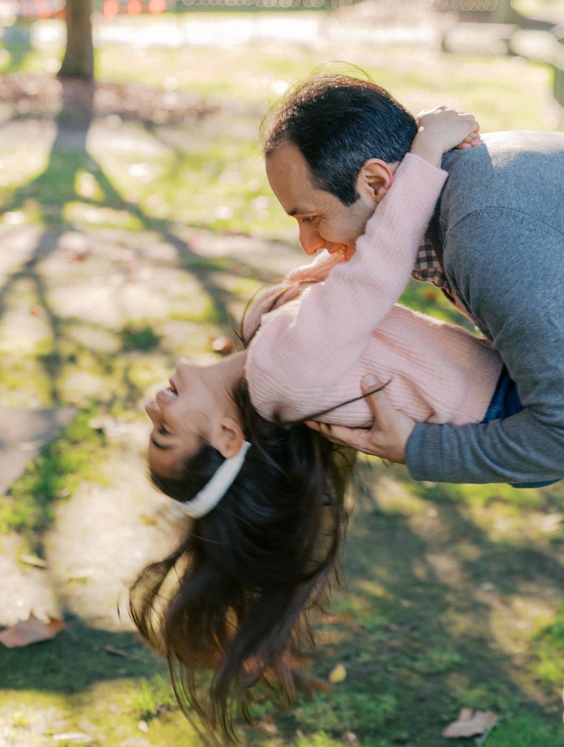 A father playfully dips his laughing child upside down outdoors in a sunny park during fall.