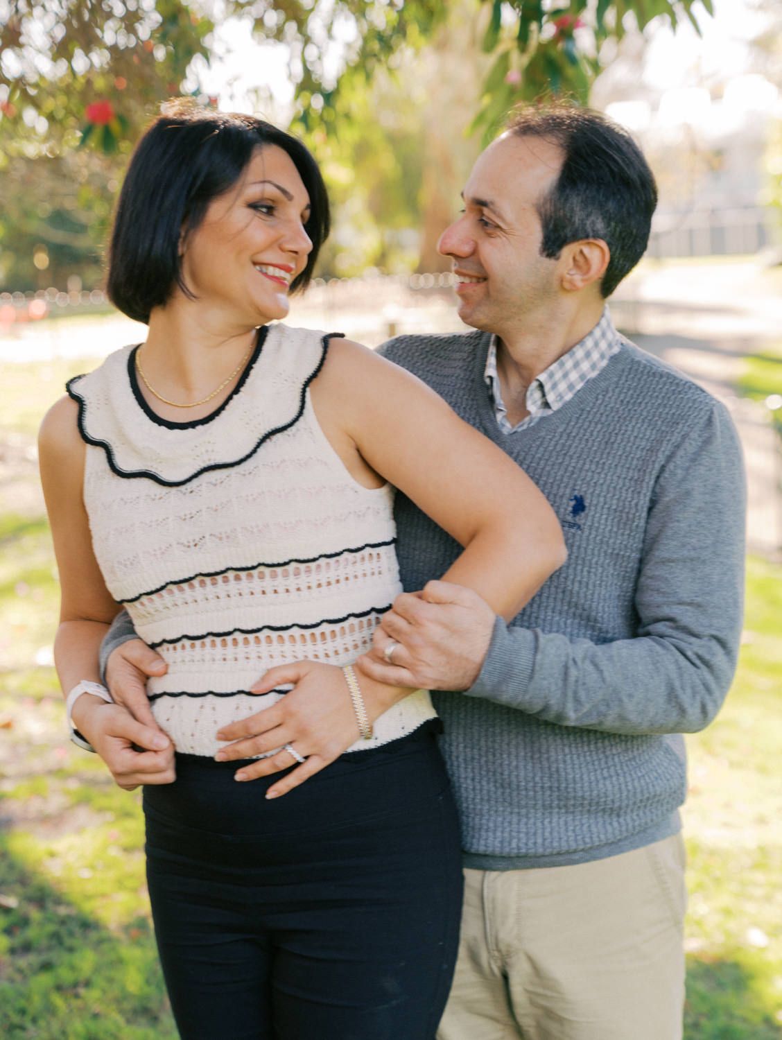 A smiling couple embraces outdoors in a park, woman in white top and black pants, man in gray sweater and khakis.