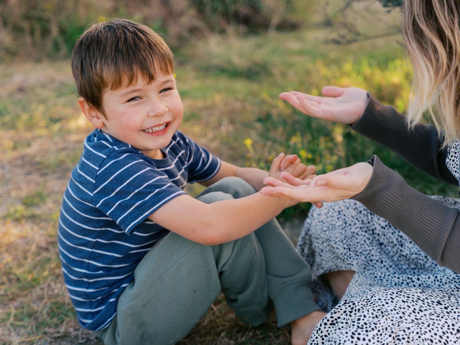 A smiling young boy sits on grass, playfully interacting with an adult's outstretched hand outdoors.