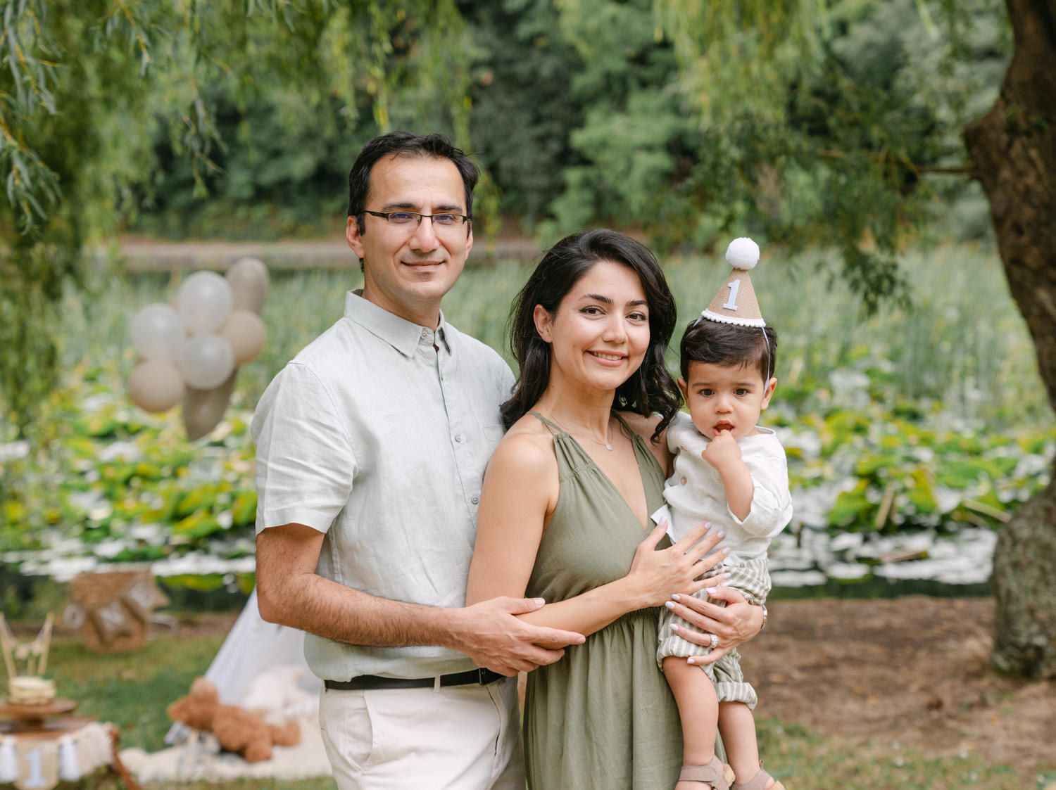 Family of three posing outdoors at a garden birthday party, parents smiling with baby wearing a party hat.