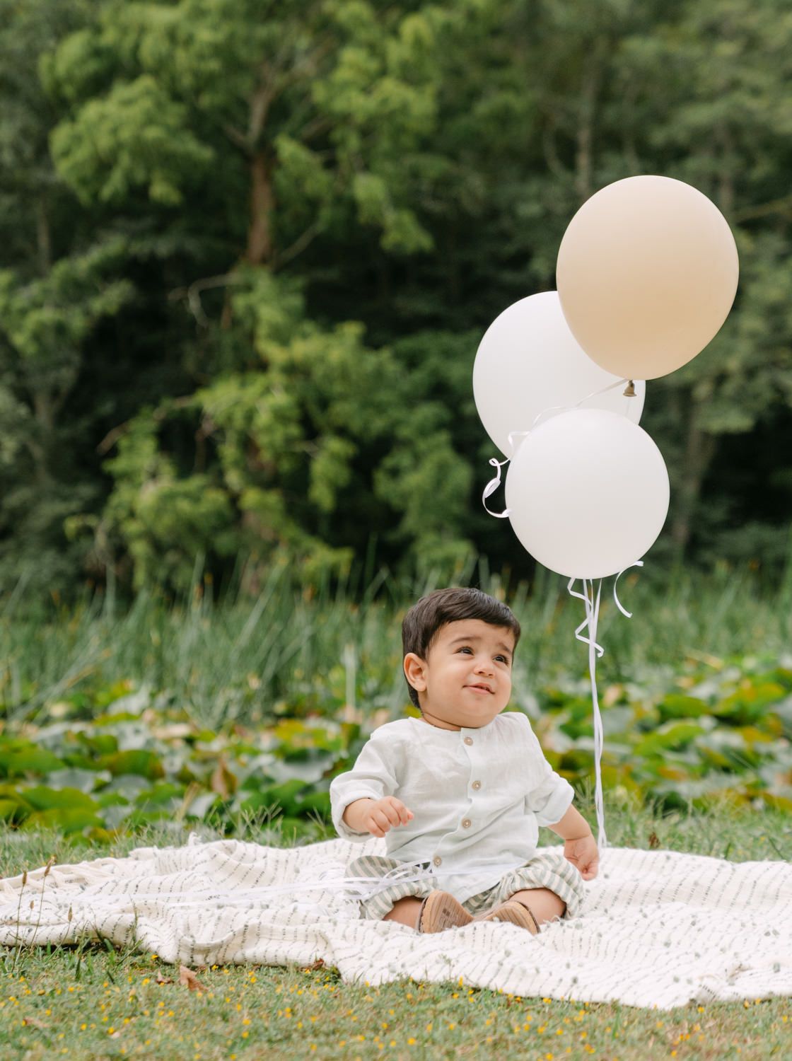A smiling baby boy sits on a white blanket outdoors holding white and beige balloons with green trees behind him.
