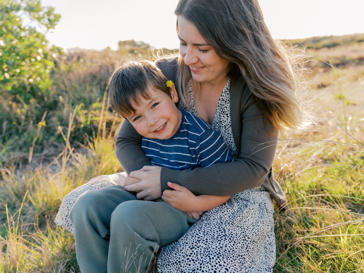 A mother hugs her smiling young son while sitting in a sunlit grassy field outdoors.