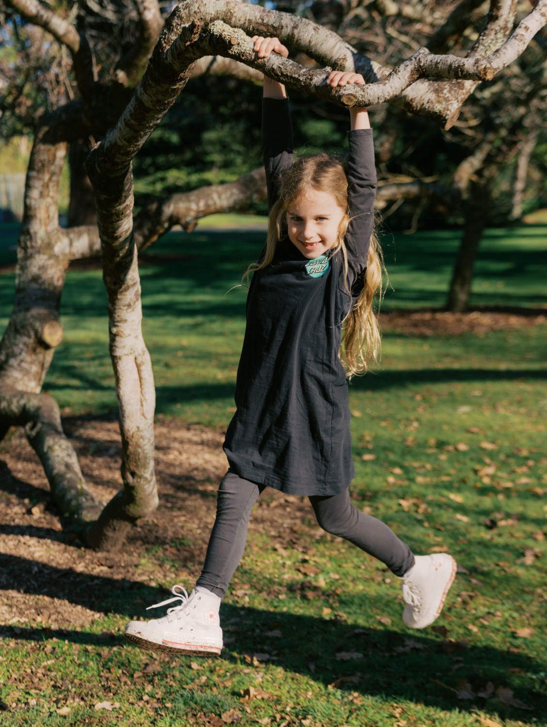 A smiling girl hangs from a tree branch in a dark dress, leggings, and white sneakers in a sunny park.