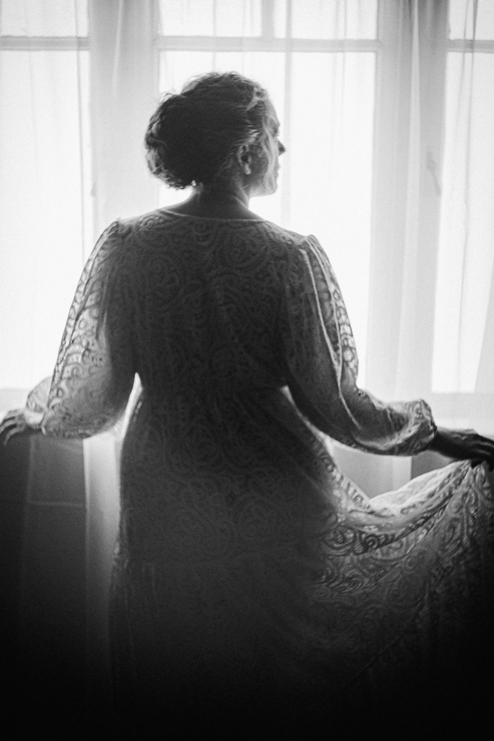 Black and white silhouette of a woman in lace dress gazing out a bright window.