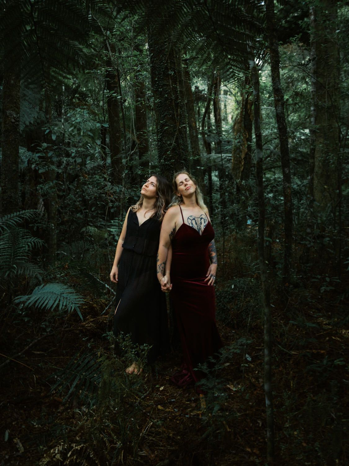 Two women in elegant dark gowns stand among lush green forest trees, gazing upward in a moody, ethereal outdoor portrait.